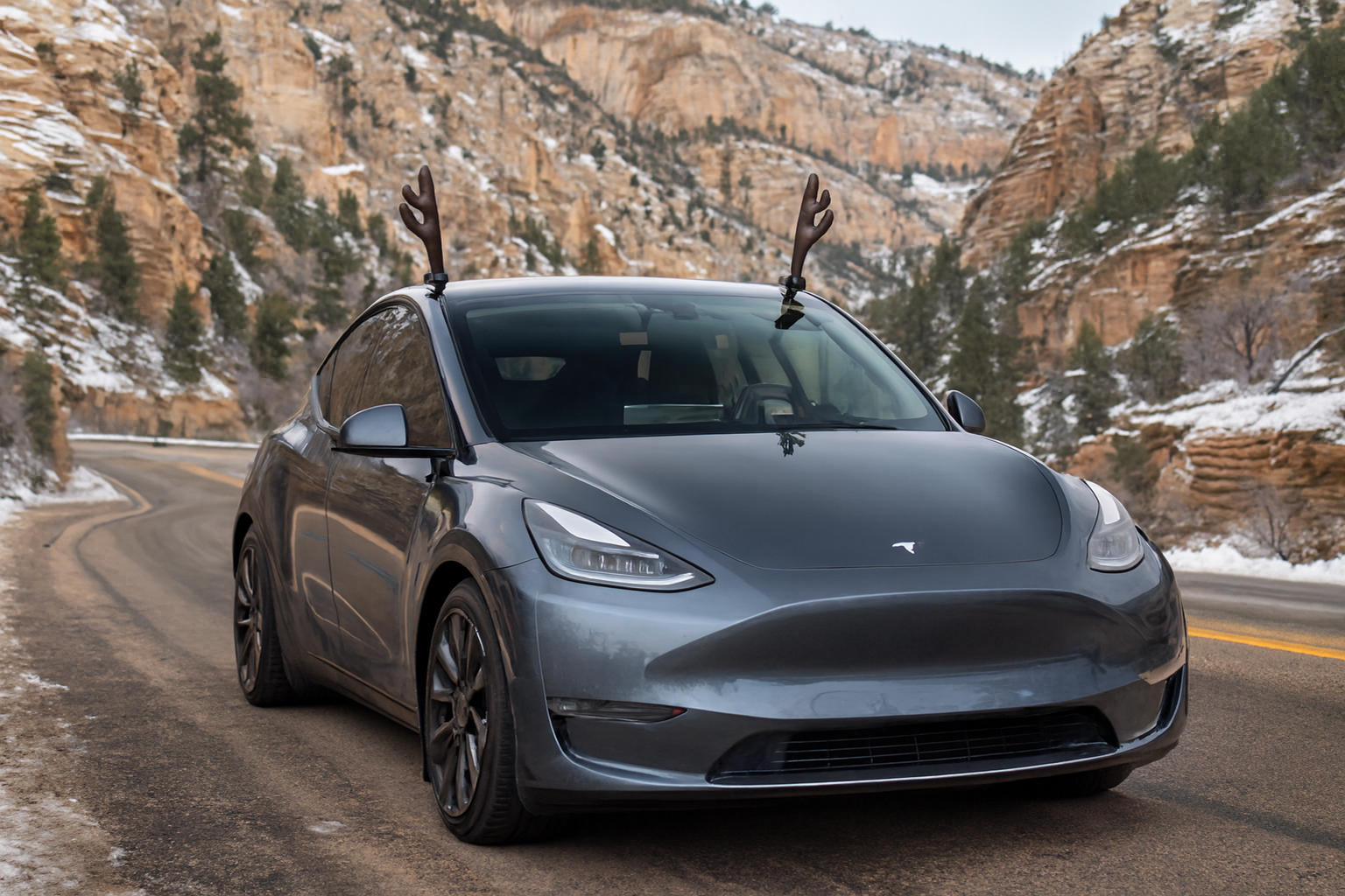Gray Tesla Model Y driving on a snowy mountain road with reindeer antlers attached to the car.